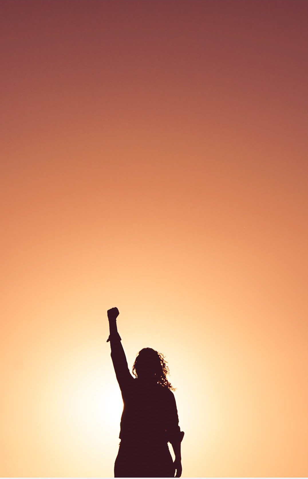A person stands in front of the sunrise and stretches his fist upwards. The person assumes a winning position.