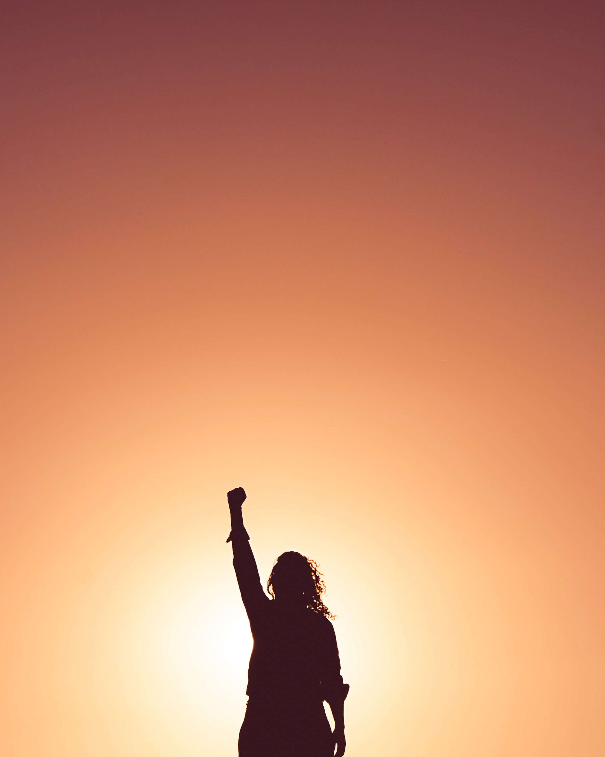 A person stands in front of the sunrise and stretches his fist upwards. The person assumes a winning position.